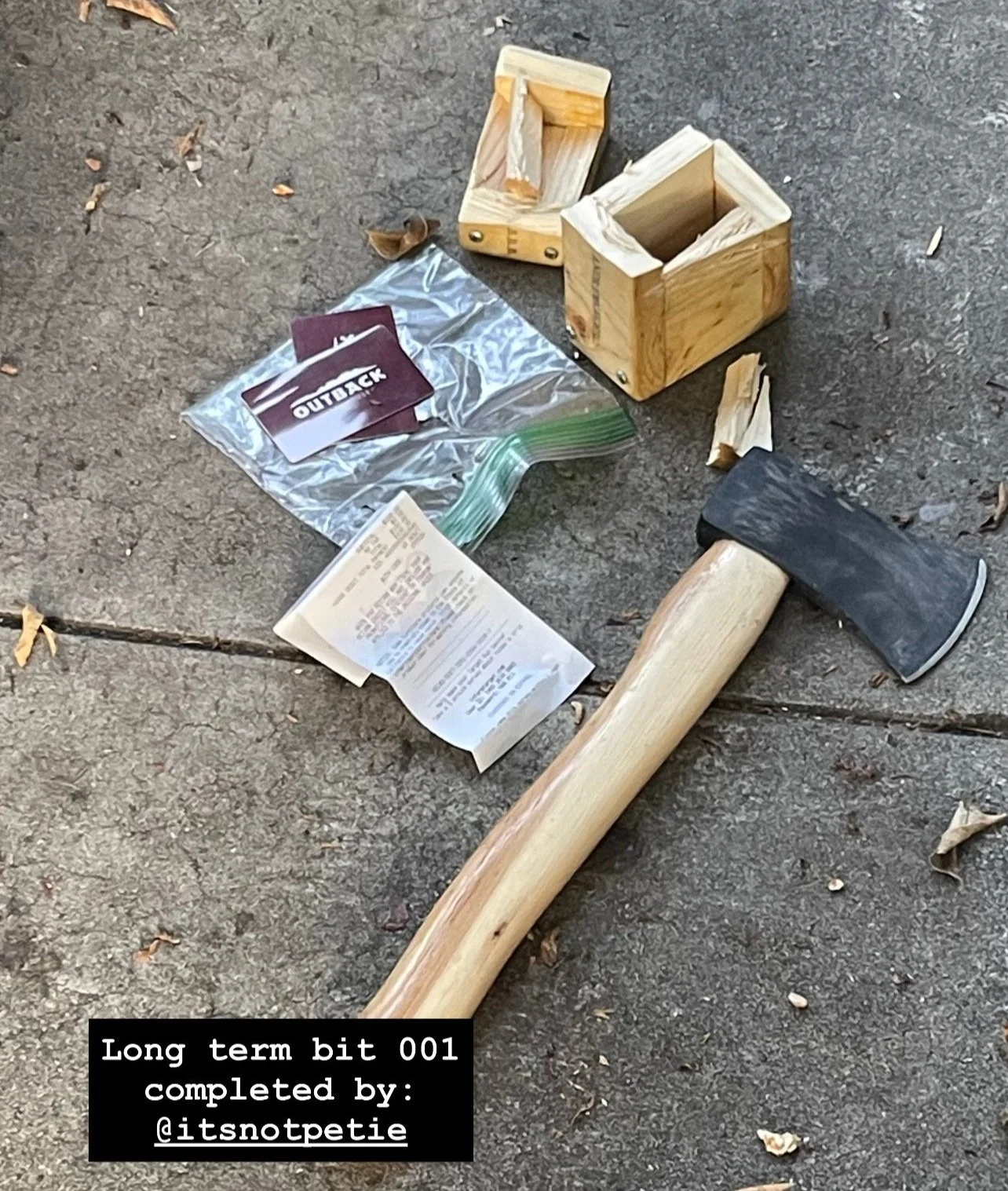 A hatchet with a wooden handle lying on a sidewalk next to a small opened wooden box, a plastic bag with Outback Steakhouse gift cards, and a receipt.