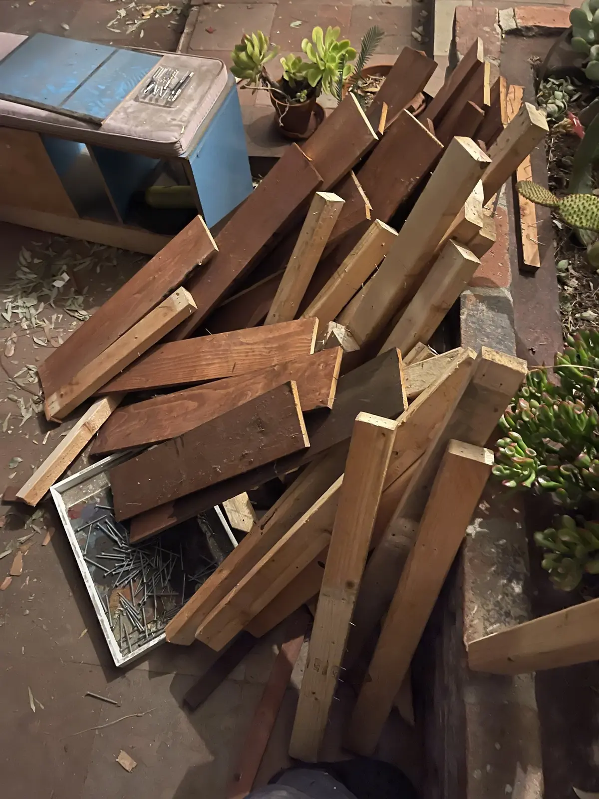 A pile of reclaimed wood boards, some stained reddish-brown, piled on a brick patio next to a tray full of pulled nails.