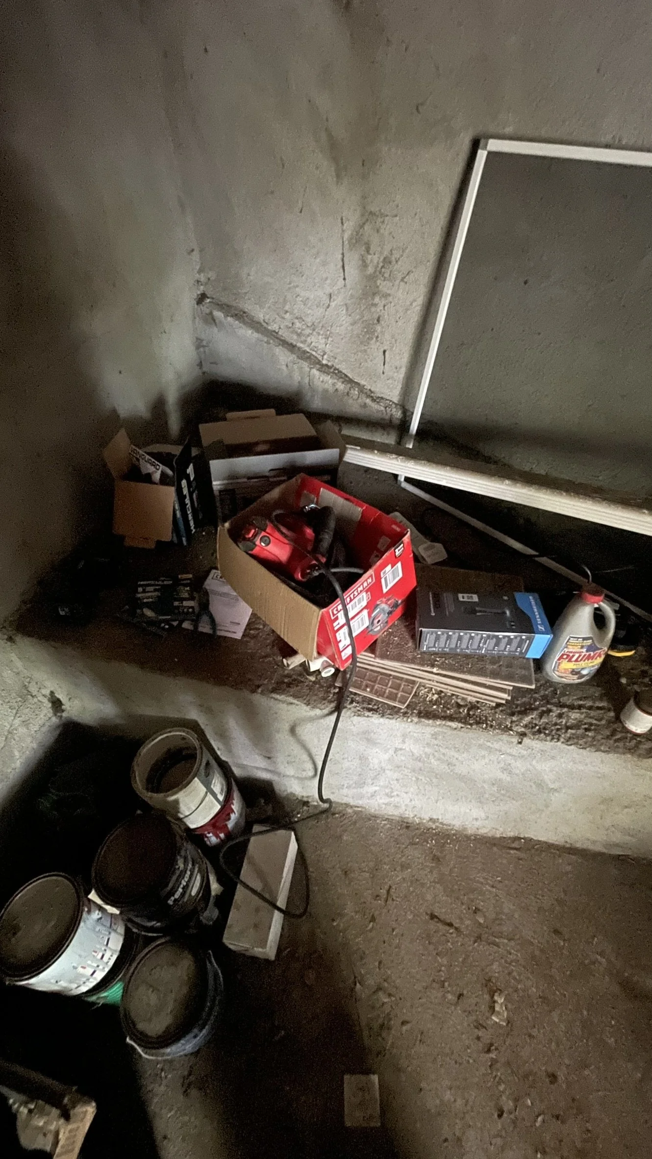A concrete storage room with a red toolbox, boxes, paint cans, and other tools stacked in the corner.