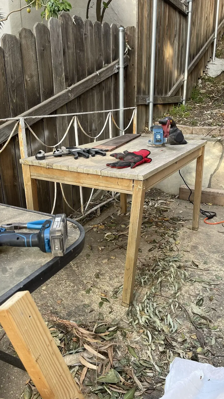 An outdoor wooden work table holding clamps, a sander, and work gloves, next to a wood fence.