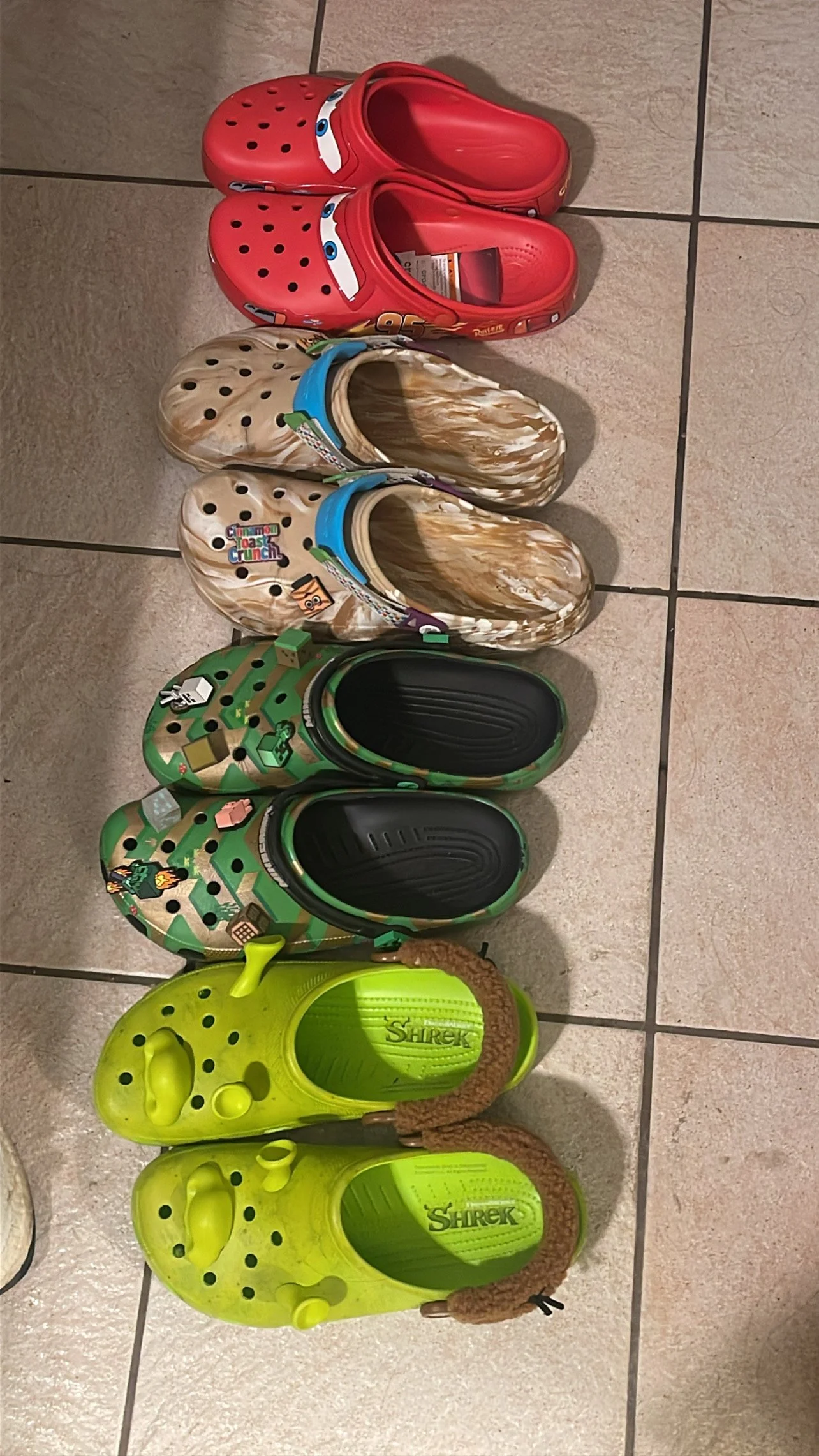 Four pairs of Crocs lined up on a tile floor: red Lightning McQueen (Cars) clogs, cream/tan marbled clogs with a Cinnamon Toast Crunch jibbitz, green Minecraft-themed clogs with black soles, and lime green Shrek clogs with an ear-shaped strap charm.